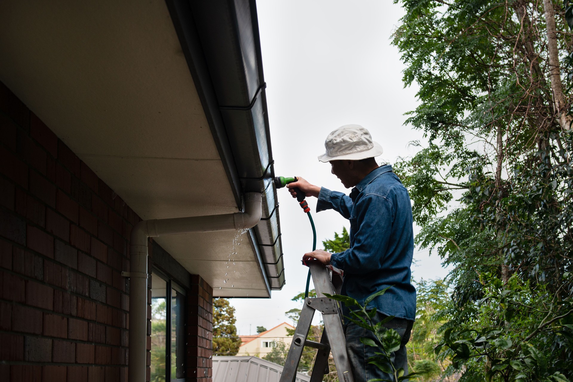 Man standing on the ladder and washing the gutter using a garden hose. Home maintenance work.